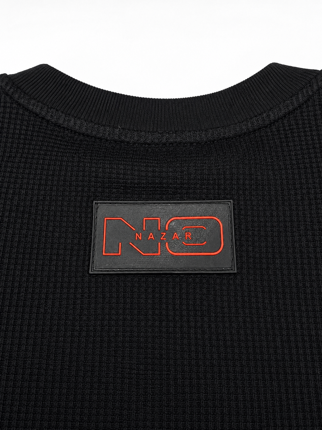 Close-up of a black ribbed knit top with a rectangular black patch on the chest featuring a red outlined 'NO' logo and the word 'NAZAR' in red.